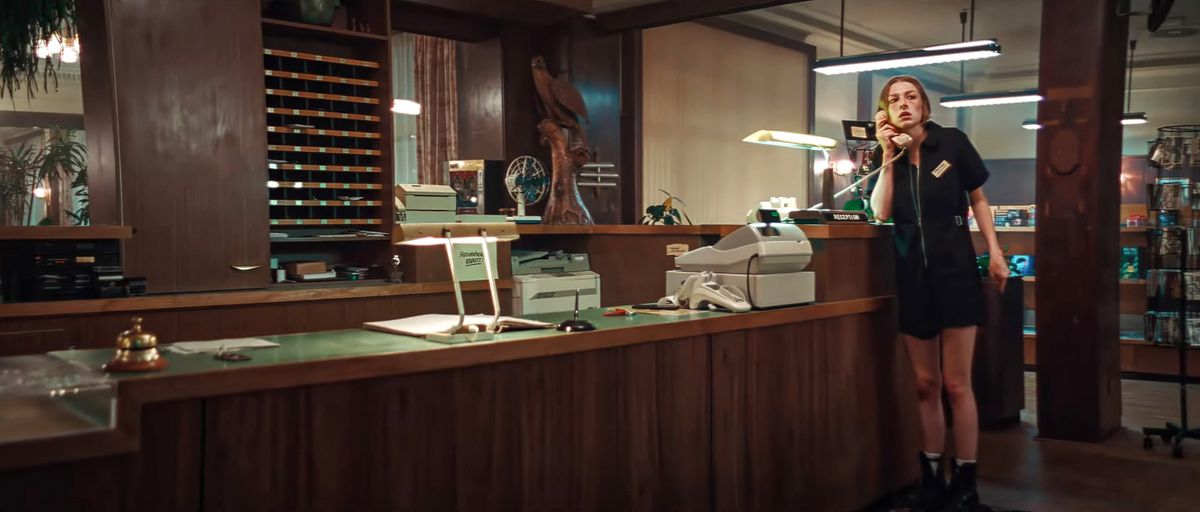 Gretchen (Hunter Schafer), a 17-year-old with a bruised face in a black thigh-length romper, stands at the check-in desk at an old resort hotel amid wood paneling and wooden sculptures in Tilman Singer’s Cuckoo