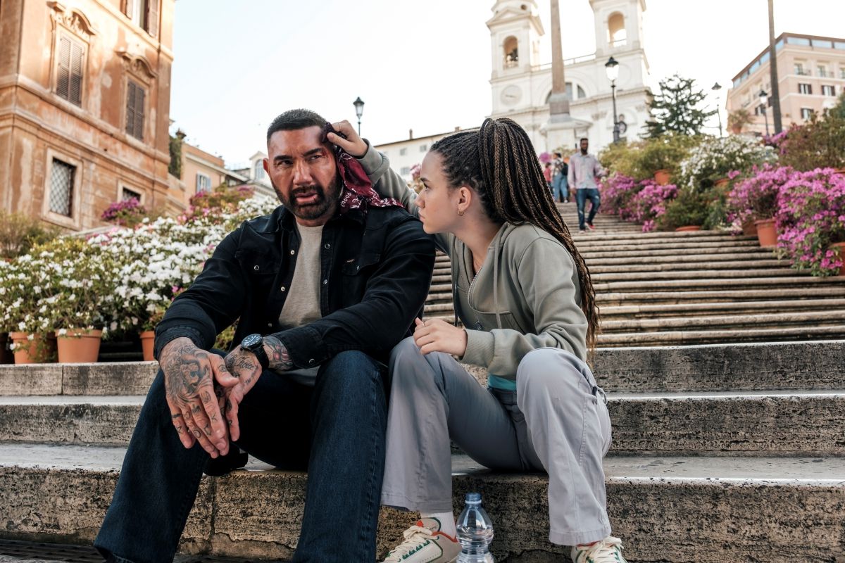 A young woman with long hair holding a handkerchief to the bleeding forehand of a large bearded man sitting on the steps of an Italian courtyard surrounded by flowers in My Spy: The Eternal City.