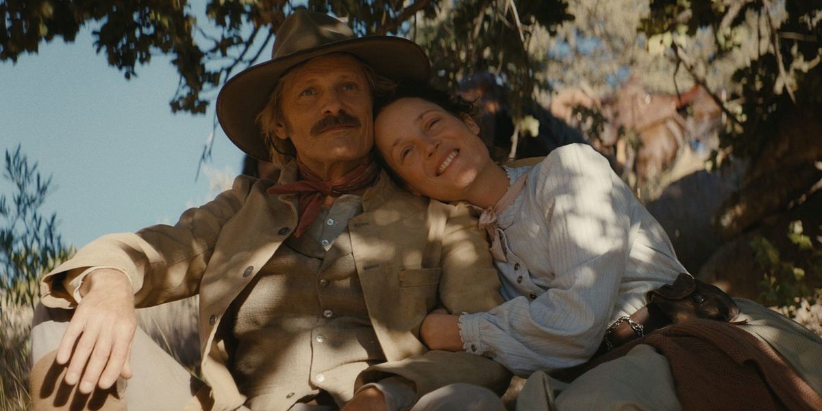 A man with a moustache wearing a cowboy hat and a tan suit sitting beside a smiling woman under the shade of a tree in The Dead Don’t Hurt.