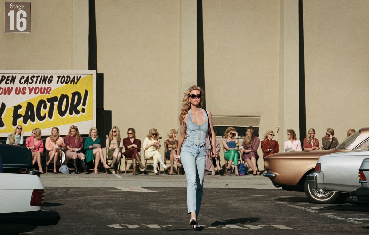 Maxine (Mia Goth) struts across a Hollywood parking lot outside a movie soundstage, with a row of other auditioners lined up in chairs behind  her, in Maxxxine 