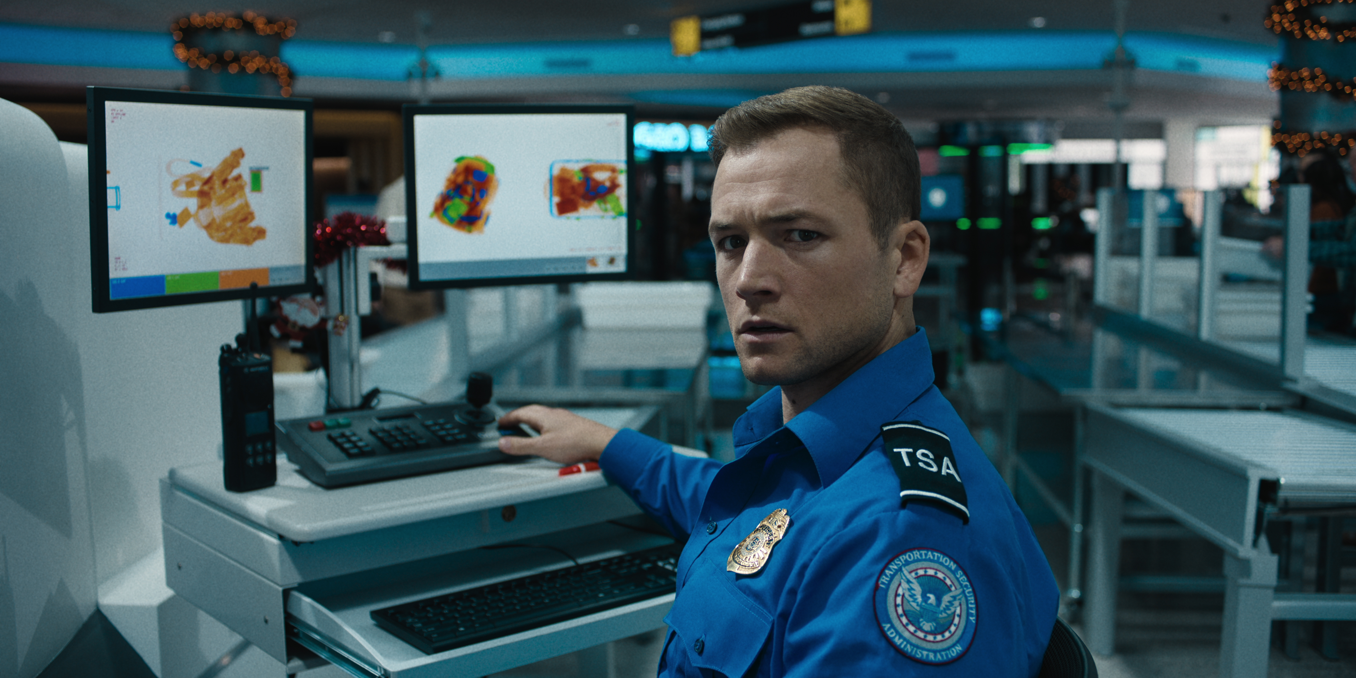 Taron Egerton dressed as a TSA agent sitting next to an X-ray console in Carry-On.