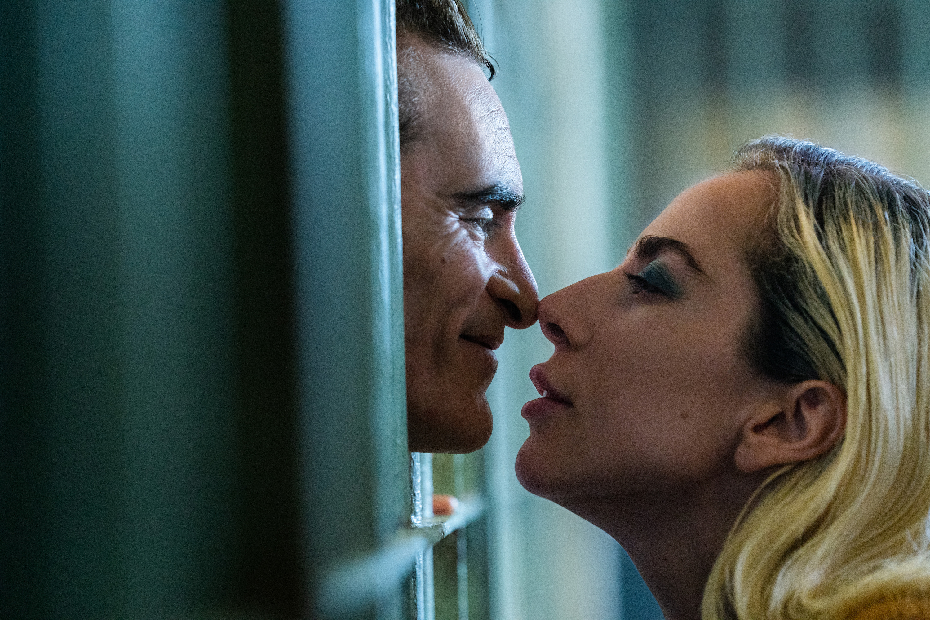 Arthur Fleck (Joaquin Phoenix) leans, smiling, between the bars of a jail cell to touch noses with his lover Harleen Quinzel (Lady Gaga) in Joker: Folie à Deux