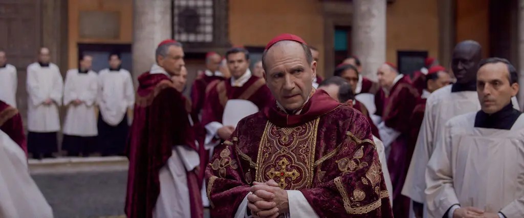 Ralph Fiennes wearing Cardinal robes in Conclave standing in the Vatican looking suspiciously at the other Cardinals around him