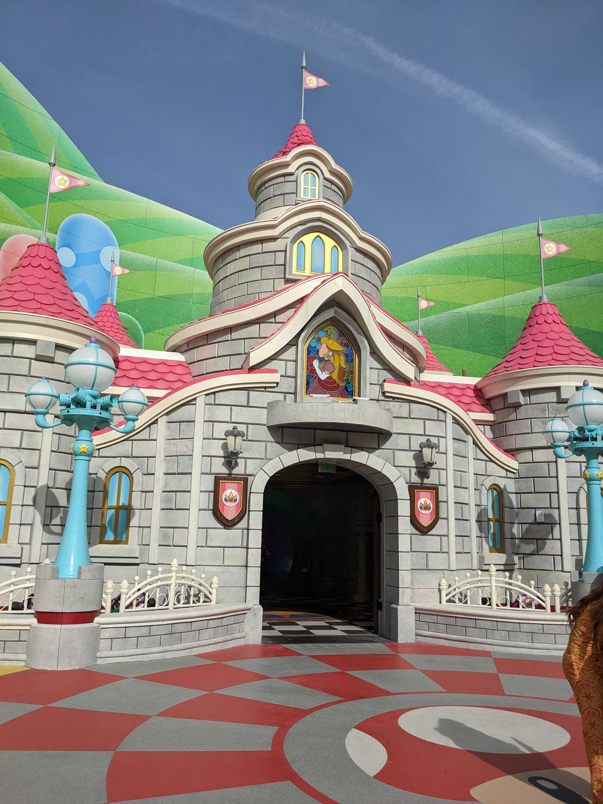 Princess Peach’s castle in Super Nintendo World. The castle is grey, has multiple stories, pink roofing, a stained glass image of a Princess Peach, and a flag on top.