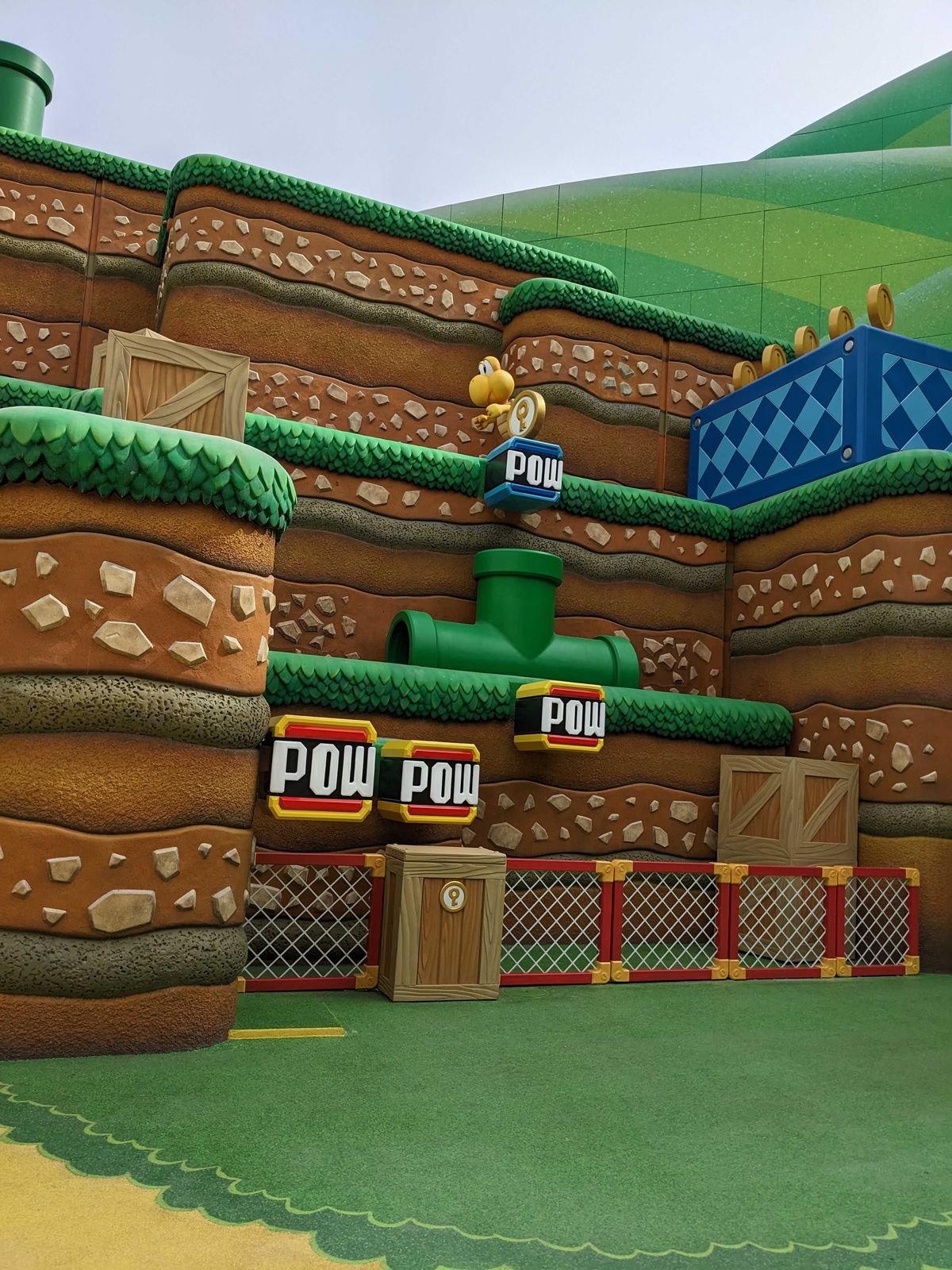 A Koopa stands above a POW button, positioned above a pipe. The activity is part of Super Nintendo World.