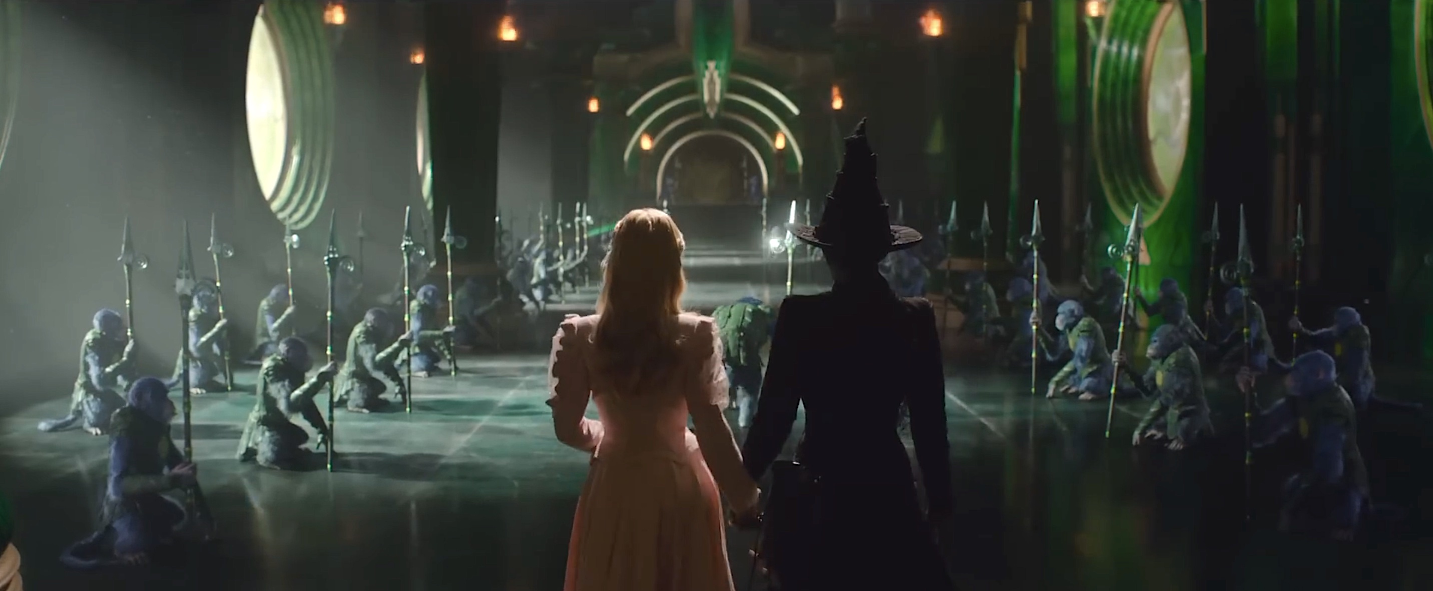 Elphaba (Cynthia Erivo, with green skin and a black witch’s hat and dress) and Glinda (Ariana Grande, all in pink) clutch each others’ hands as they look down a dark corridor lined with blue-furred, armored, squatting baboon soldiers holding pikes in Wicked: Part I